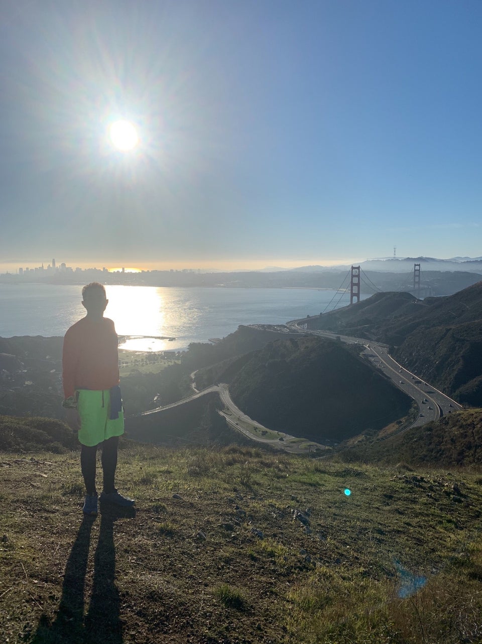 Tantek standing on a hill, backlit by a bright sun in the blue sky, the San Francisco skyline and bay behind him, Golden Gate bridge towers visible behind the Marin headlands to the right, the 101 freeway far below, sparse with tiny cars.