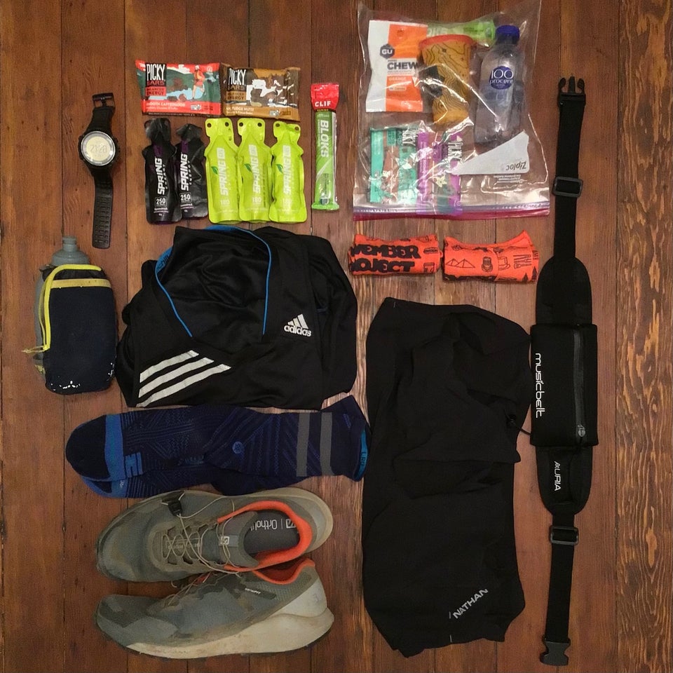 Trail running shoes, socks, shorts, singlet, water bottle, Suunto watch, Spring Energy gel packets, Picky bars, November Project buffs, ziplock with drop bag supplies, and waist belt arranged orderly on hardwood floor.