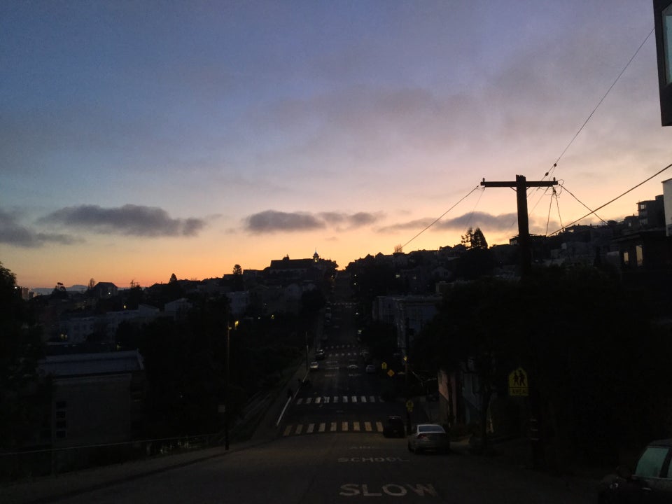 Looking down a street in Potrero hill that dips down and goes back up, at dawn with a purple blue sky that turns orange at the horizon, backlighting the outline of buildings and a telephone pole.