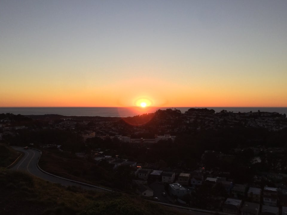 Sun half set just above the Pacific Ocean horizon, backlit dark trees on hills in the distance below, scattered houses on the hills in the shadow, a winding road nearby on the side of the hill below.