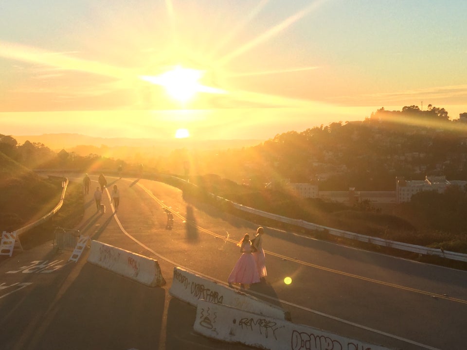 Very bright sun emitting lots of sunbeams barely over the Pacific Ocean horizon, lighting up pedestrians along the road, including two women dressed in pink dresses.