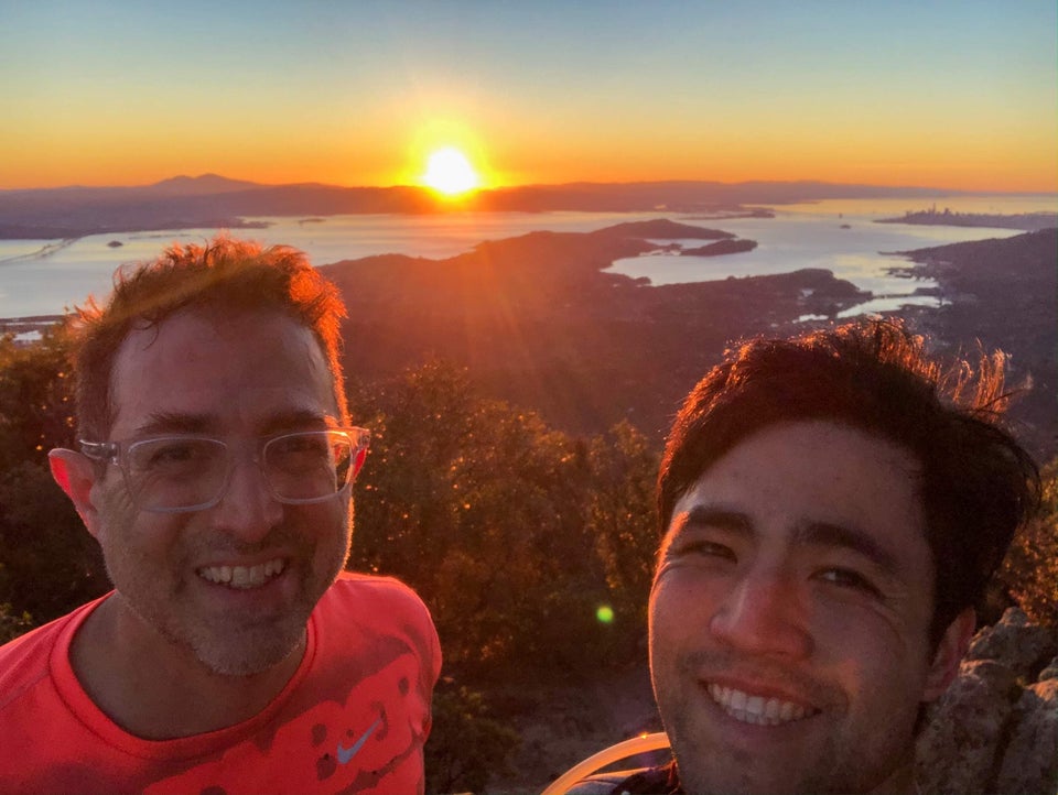 Tantek and Bryan being backlit by the sun just rising above East Bay mountains, the bay far below, along with the Tiburon Peninsula, a tiny Bay Bridge and San Francisco skyline, and nearby bushes behind Tantek and Bryan.