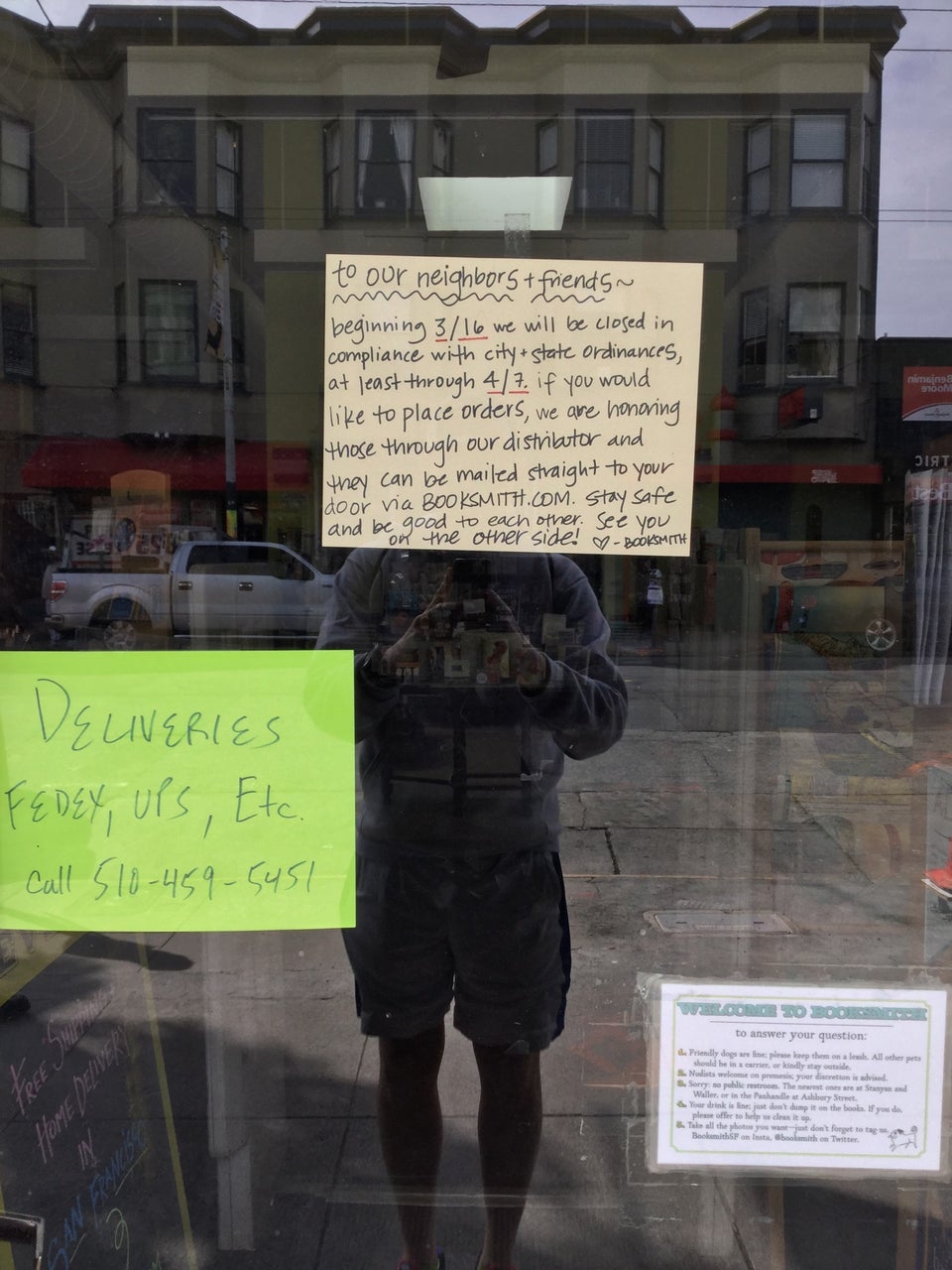 Handwritten sign outside Booksmith with instructions for ordering online, and another sign with instructions for deliveries on the glass door,  which itself reflects the photographer taking the photo, and the street & building behind him.