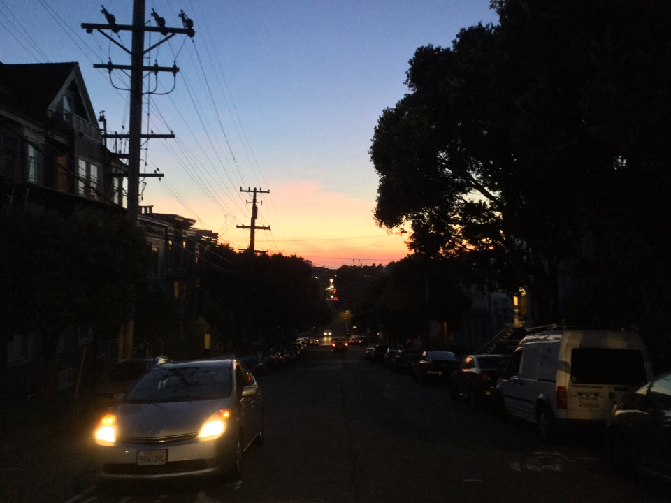 Sunset in the distance over the ocean, buildings on both sides, a car coming up an uphill street with its headlamps on.