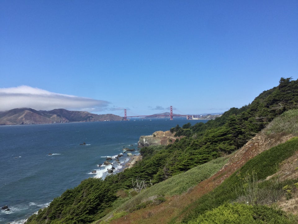 Golden Gate Bridge in the distance under blue skies, Lands End hills in the foreground, and a cloudbank hovering over the Marin Headleands on the left.