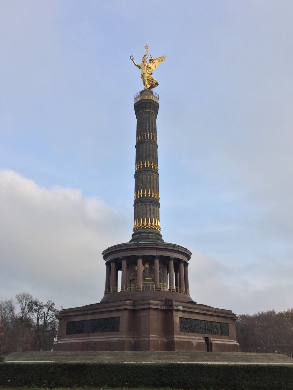 Berlin Victory Column under a partyly cloudy sky, sitting atop its base, with trees in the background on both sides.