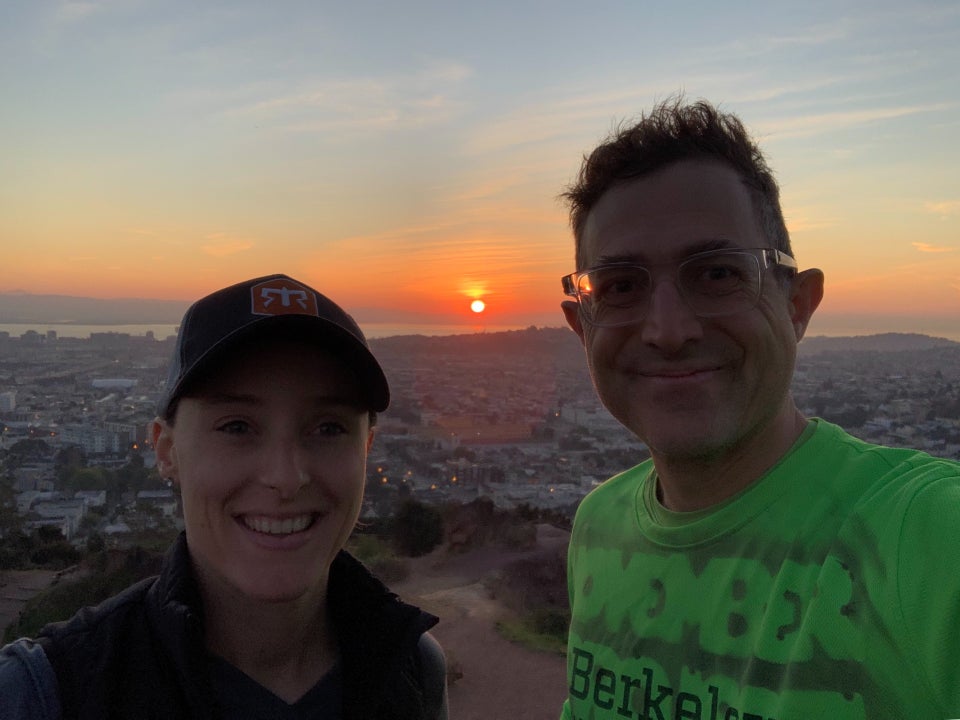 Brooke and Tantek with the sunrise behind them, the sun glowing red as it’s filtered through some low hanging haze just above the East Bay Hills, a sliver of bay visibe below it, then distant San Francisco hills, buildings, finaly trees and trails of Corona Heights Park below.