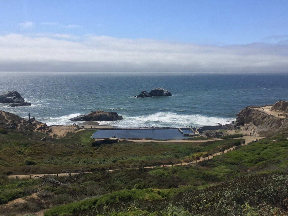 Sutro baths at the Lands End Lookout with green trails in the foreground, and a blue Pacific Ocean in the background, fog gathering in the distance.