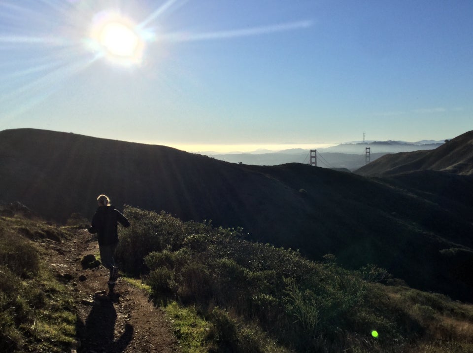 Emily runnin on Alta Trail, bright sun overhead in a clear sky, Marin Headlands in front of us, the tops of the Golden Gate bridge towers peeking up behind them, a tiny Sutro tower in the distance.