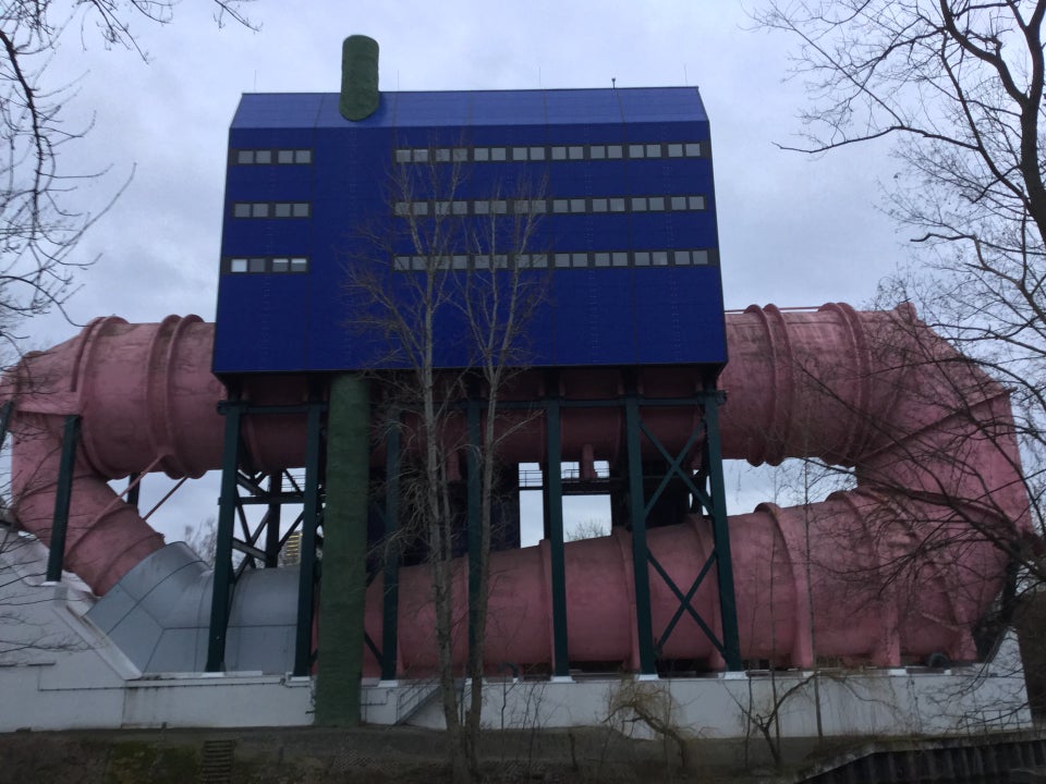 Gray overcast sky, rectangular blue building with three rows of small square windows, on stilts above the shore of the canal, a giant pink pipe emerging from the side of the building, going underneath, and re-entering on the other side.