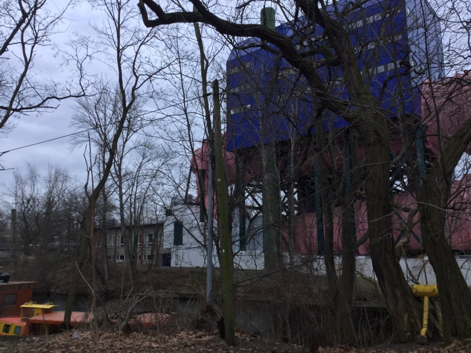 Blue building on stilts and a giant pink pipe in the background on the other side of a canal, partly hidden by a cluster of leafless trees in the foreground, a orange & yellow colored houseboat on this side of the canal, and what looks like a giant yellow plastic mallet leaning against a tree on the lower right.