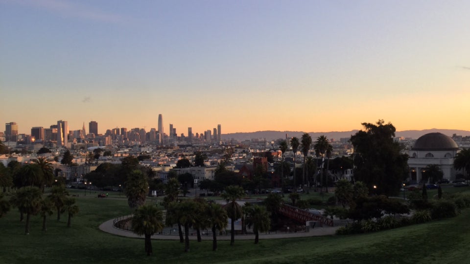San Francisco skyline and East Bay hills bathed in sunrise on the horizon, Dolores park spanning the foreground with palm trees, and walking paths curving through the lawn.