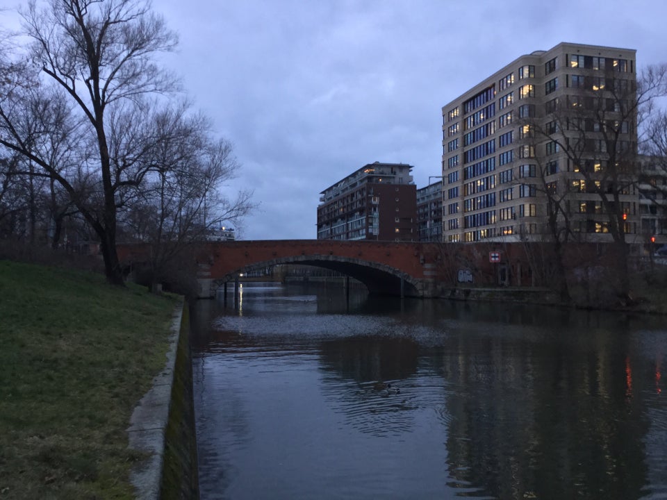 Early evening overcast cloudy gray sky over buildings on either side of the red Dovebrücke bridge, green lawn on the left bank with a bare tree, a few bare trees on the right side, not much reflected in the canal below.