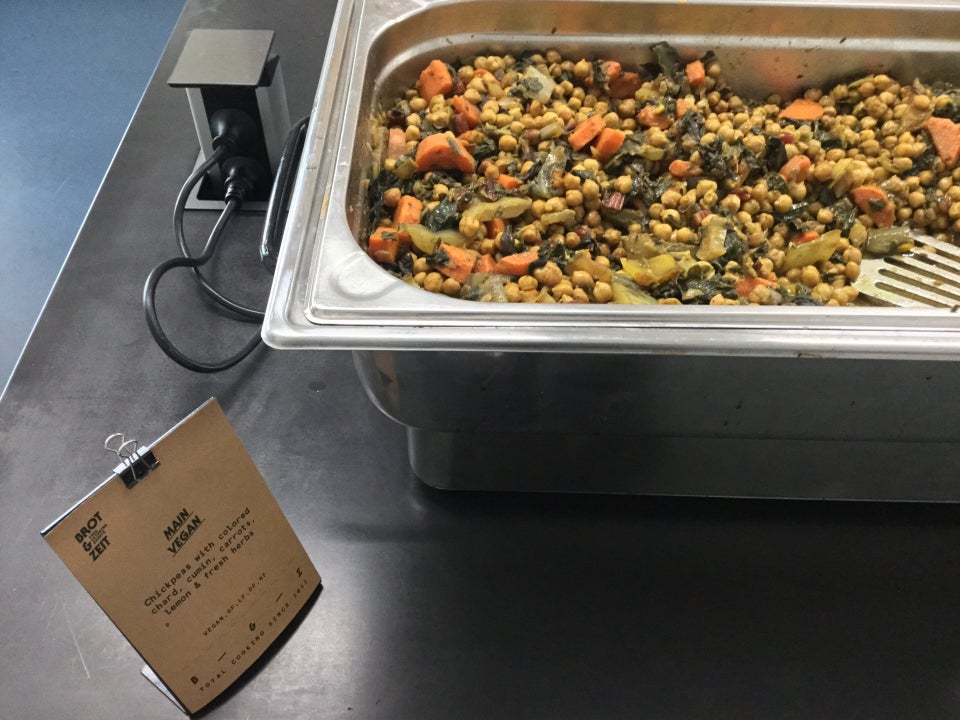 Vegan catered food: chickpeas with colored chard and other vegetables in a large metal container, being heated electrically, on a black table, with a blue floor below.