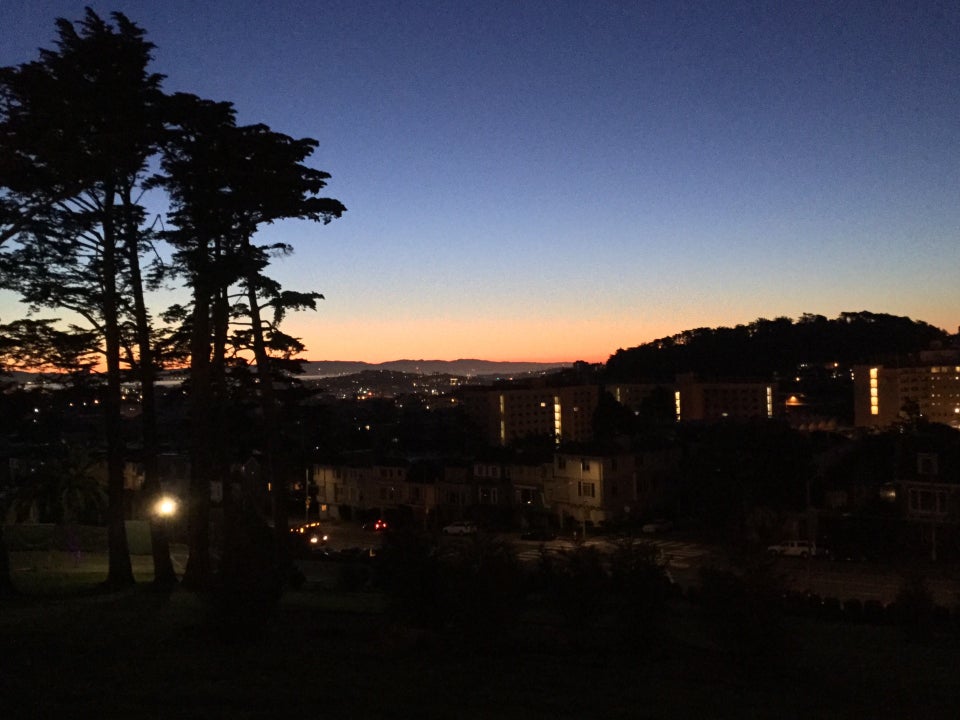 Blue to light blue gradient over a bright orange band on the horizon, dark backlit trees in the foreground, and buildings in darkness with their lights on.