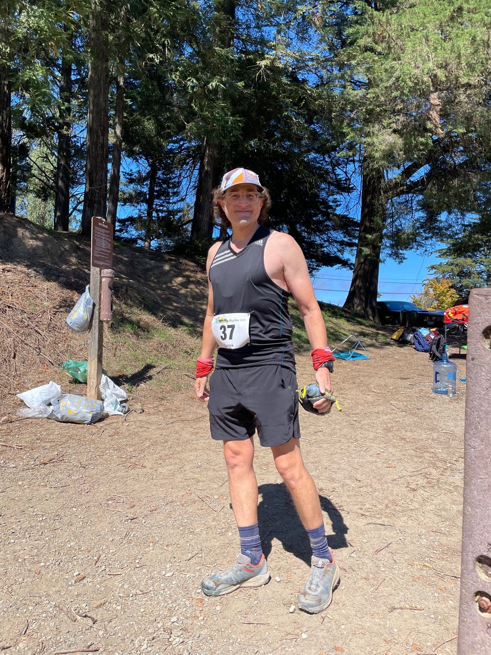 Tantek standing at an angle on a wide trail in bright sunshine with pine trees behind him, holding his water bottle, leaning forward on his right foot, head turned slightly to face the camera and smile.