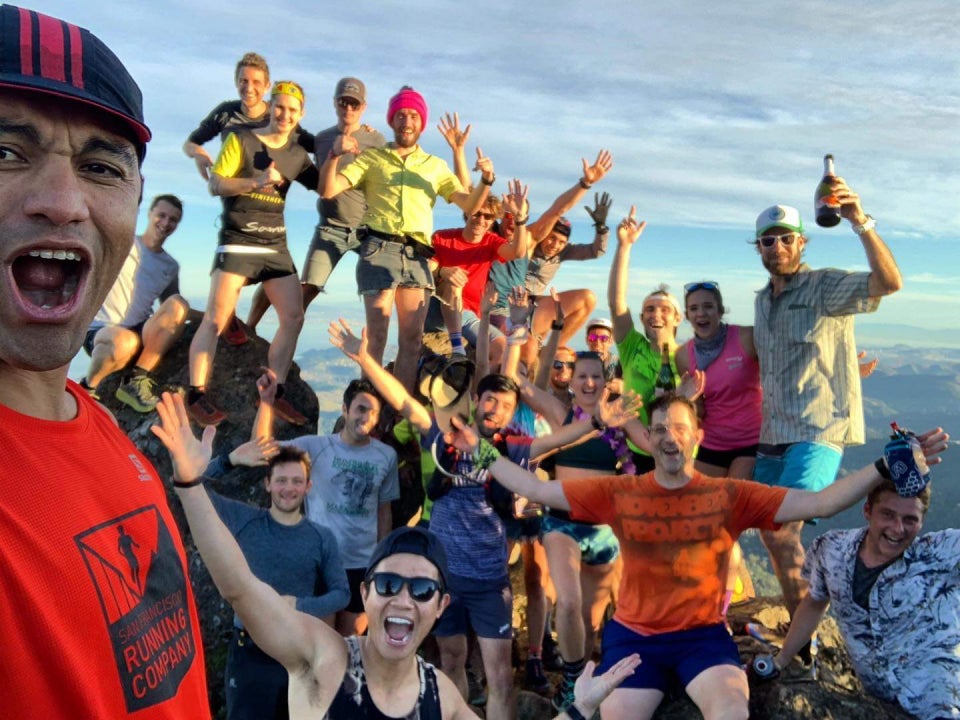 Huge crowd of trail runners cheering with their arms up, perched on top of rocks at the Mount Tamalpais East Summit with the bay and lands beyond in the background.