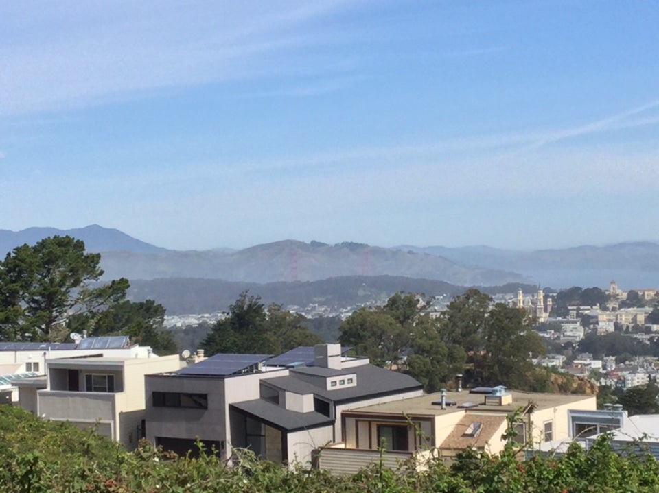 View of Mount Tam in the distance, Marin Headlands in front of it, the Golden Gate Bridge, the Presidio hills, trees close by, and the roofs of houses near by, some with solar panels on top.