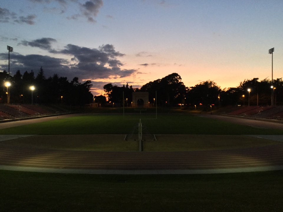 Just after sunset at Kezar stadium,mostly clear sky with gradients from light blue to orange and yellow, above dark backlist trees, the few minor stadium lights lighting up parts of the track and a bit of the center lawn, looking down the field from one field goal to the other with the orange sky in the background.