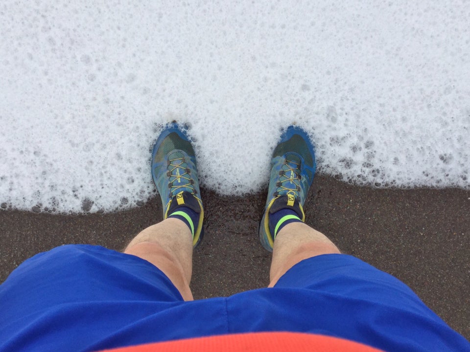 Looking down at the surf coming up the sandy beach and surrounding trail running shoes, socks, legs, and shorts above them.