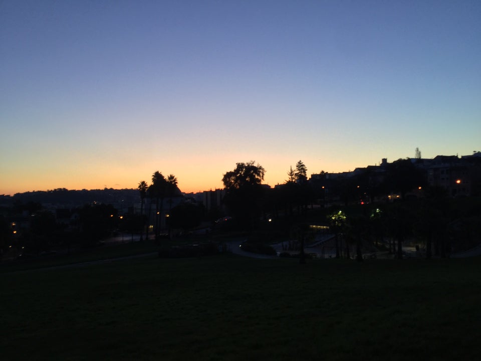 Medium blue clear sky above an orange band at the horizon, backlighting trees above Dolores Park still in darkness, yet just light enough to see a dark green tint of the grassy field.