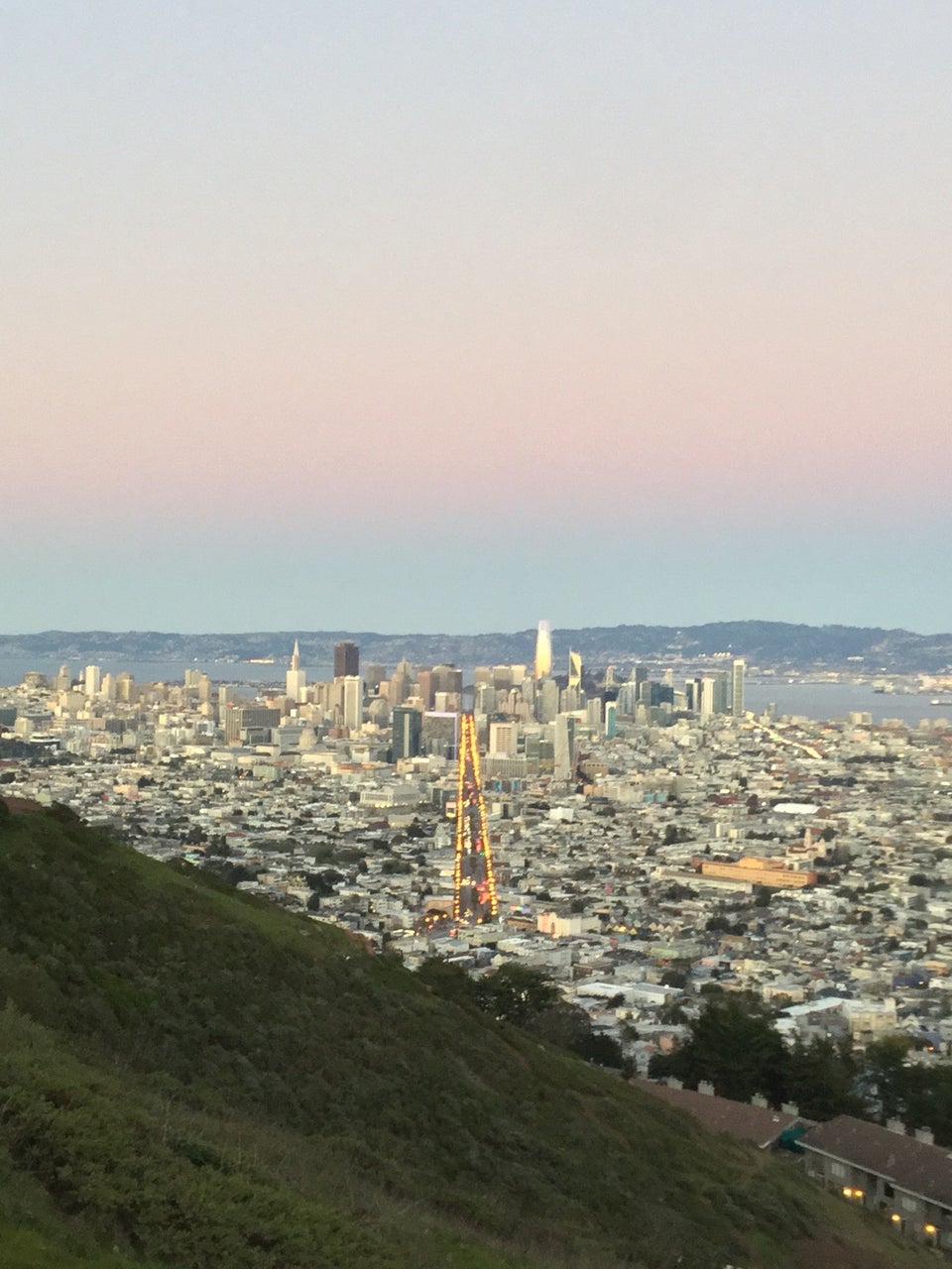 Clear gradient of white to yellow, orange, pink, purple, and blue sky above East Bay hills in the distance behind the San Francisco skyline, buildings spreading outward on either side of Market street lined with lit street lamps, nearby green hillside in the foreground.