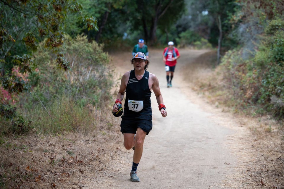 Tantek running with a focused look in a 2020 MUC50 cap, black singlet & shorts, on a dirt trail with grass, bushes, & trees on both sides, and two runners out of focus ~20 meters behind him.