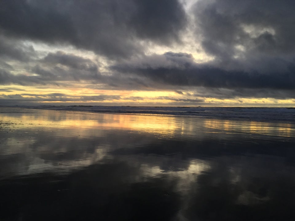 Dark clouds with wisps of sky barely visible between them, one of them making a tilted V, over a yellow orange horizon from the recent sunset behind the Pacific Ocean, waves crashing, leaving a smooth wet sandy shore with a mirror image reflection of the bright horizon, and a negative space letter T from the reflected wisps of sky surrounded by dark clouds