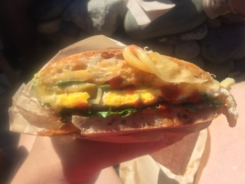 A hand holding half a ciabatta breakfast sandwich in its half-wrapper, under sunlight bright enough to give it all a soft glow