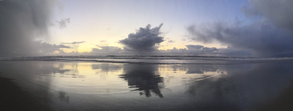 Panoramic view of storm clouds to the South, an opening of nearly clear sky with clouds on the horizon and one big cloud nearer, and more storm clouds to the North above the Pacific Ocean at Ocean Beach with waves crashing and leaving behind a wet reflection on the sand of the same clouds but darker and textured with ripples as the water recedes back to the ocean.