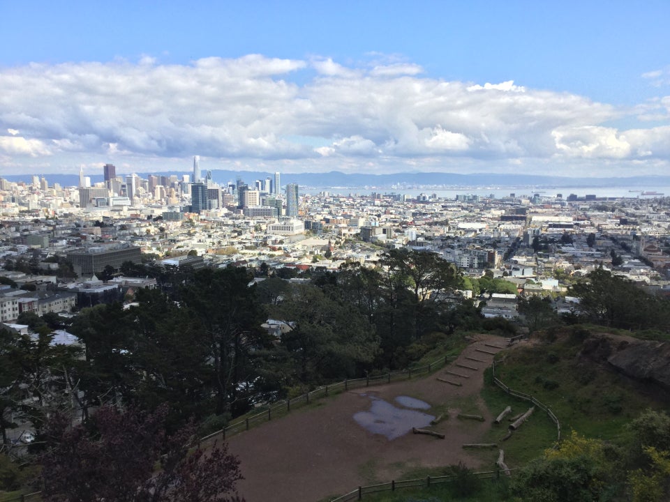 Light blue skies, low hanging scattered white gray clouds over East Bay hills in the distance, downtown San Francisco, and trees, green bushes, grass, & dirt paths of Corona Heights Park below, with a couple of large puddles from recent rains.