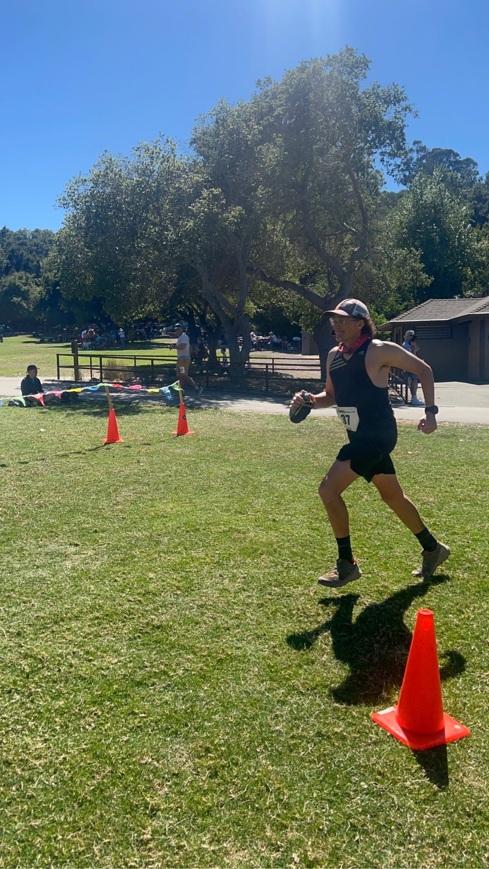 Tantek running mid-stride, arms swinging, on a lawn between orange cones with a park building and trees behind him.
