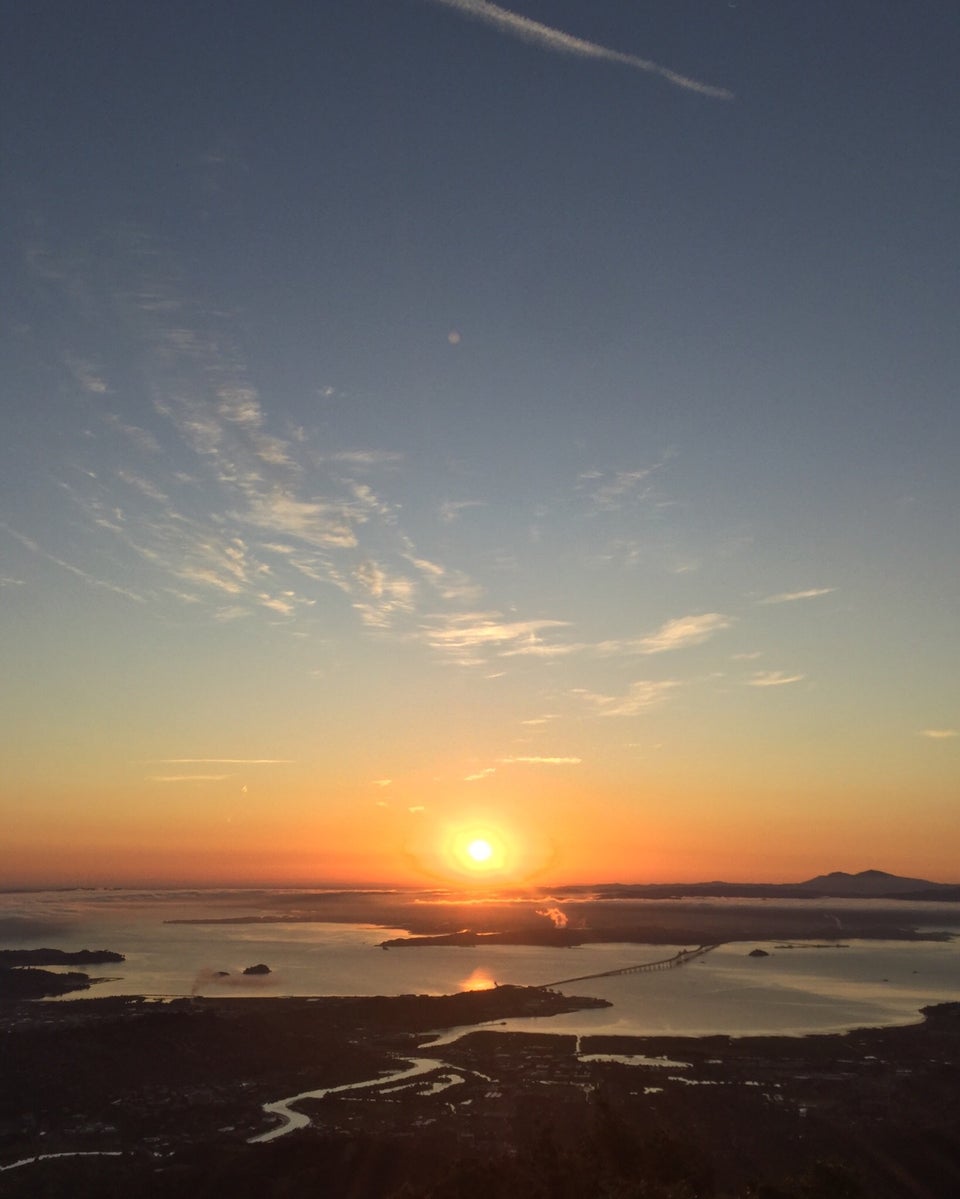 The sun rising just above East Bay hills under an orange to light blue sky as viewed from the East Peak of Mt. Tam