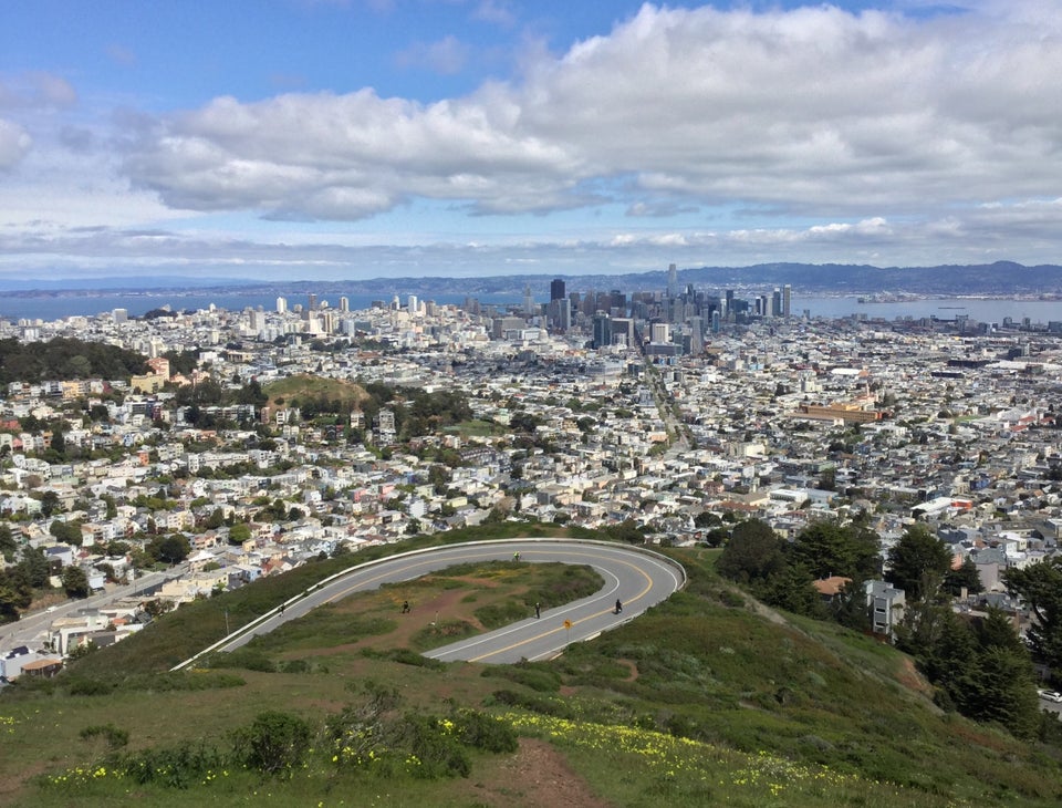 White clouds in a blue sky above distant East Bay hills, clearly visible, San Francisco downtown, looking down Market street, and the nearer neighborhoods, just behind the hairpin turn and Twin Peaks hill below.