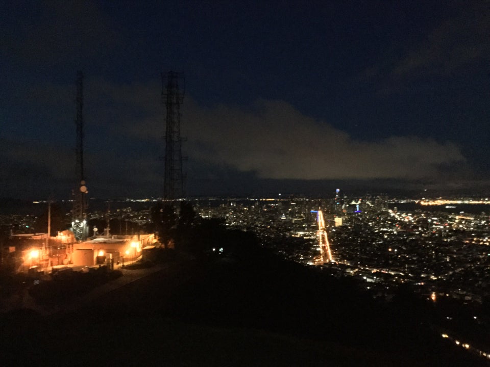 A large white cloud on a very dark blue sky underlit by city lights, most prominently two segments from streetlights outlining the edges of Market street converging in the distance, a dark hill in the foreground, to the left the top of Twin Peaks and its few utility buildings lit with a few lights, two thin dark transmission towers rising above them.