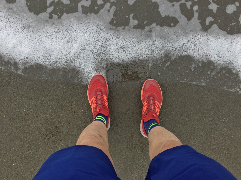 Looking down shorts and trail shoes standing on the beach, the surf receding