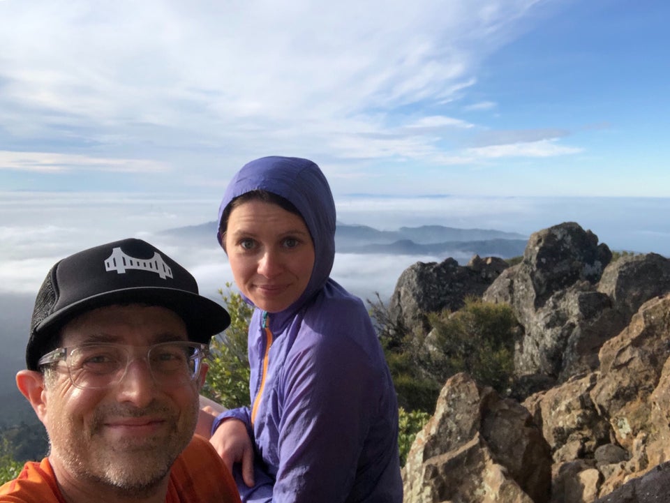 Tantek and Hannah sitting on top of Mt. Tam, bushes and rocks right behind them, clouds and hills below in the distance, partially visible San Francisco building tops poking through thick fog in the far distance