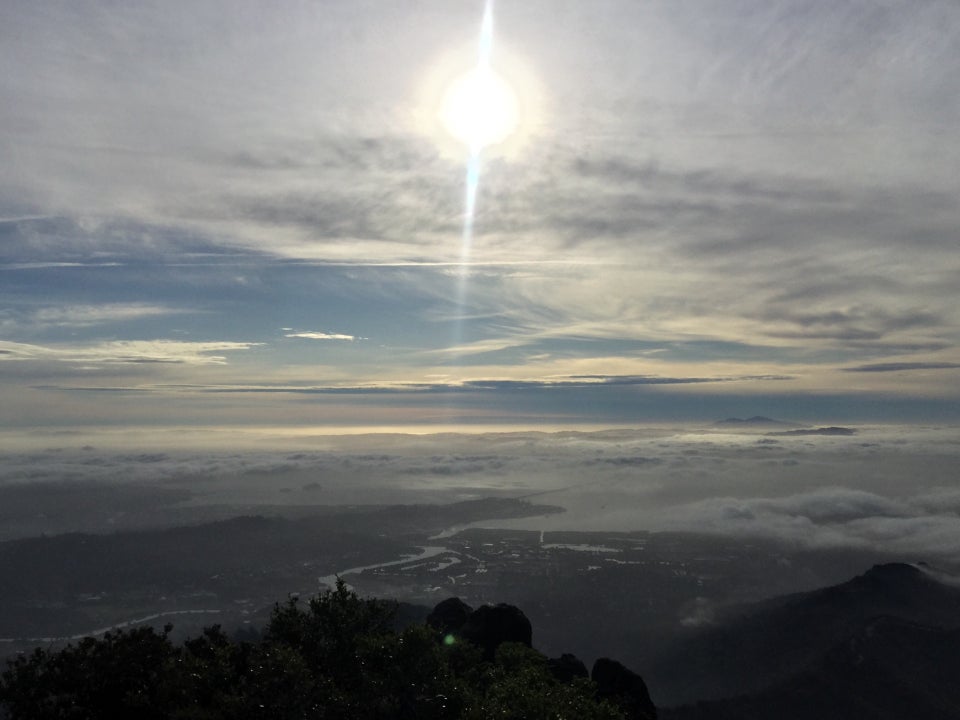 The sun shining through an upper cloud layer, high above distant mountain peaks, themselves poking through lower clouds, a partially visible bay, and nearer hills, bushes and a rocky outcropping in the foreground